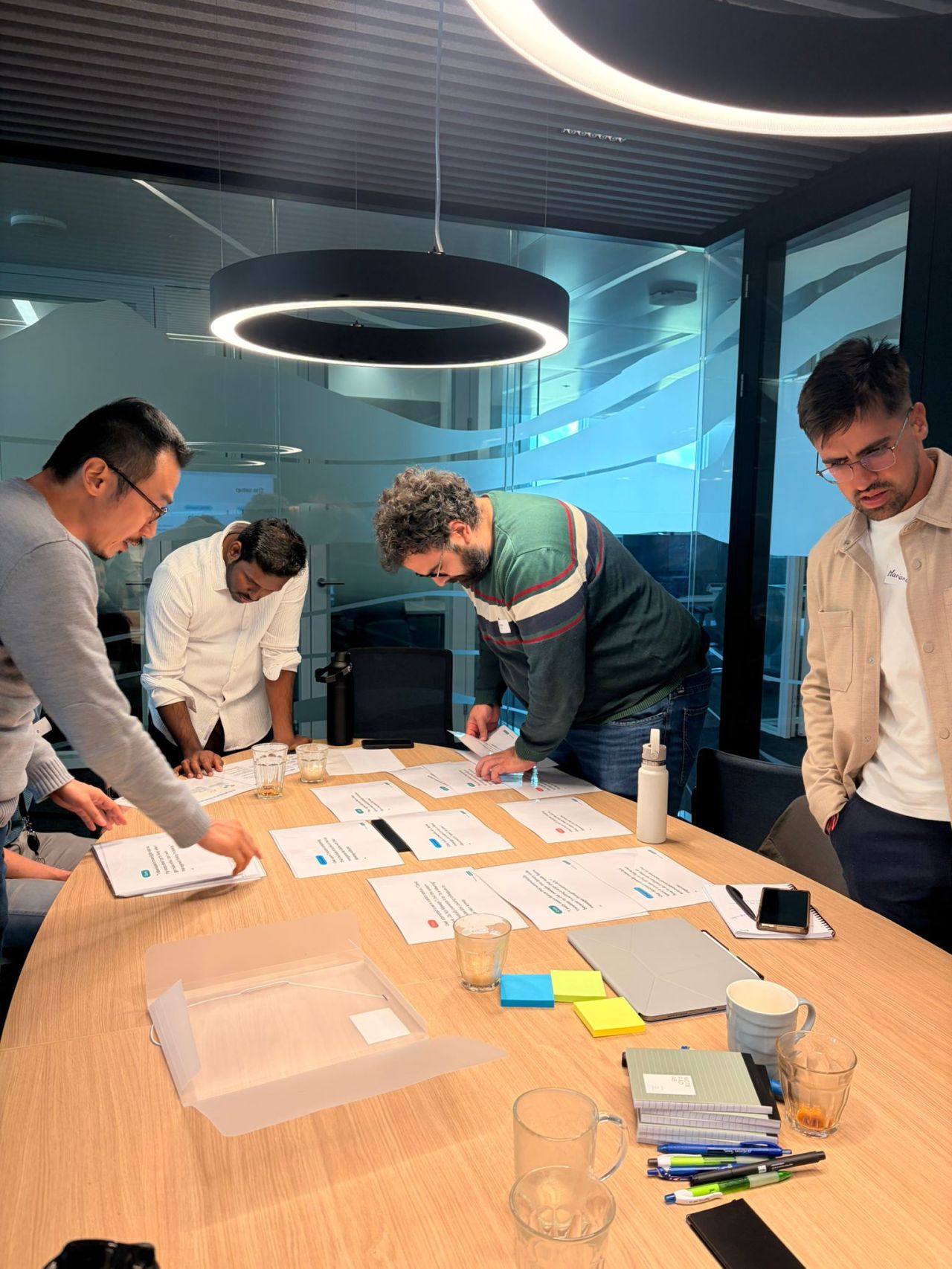 Four people standing around a meeting room table covered in printed sheets of research data, leaning over it and moving the sheets into groups. Glasses, notebooks and a tablet have been pushed to the edges of the table to make room.