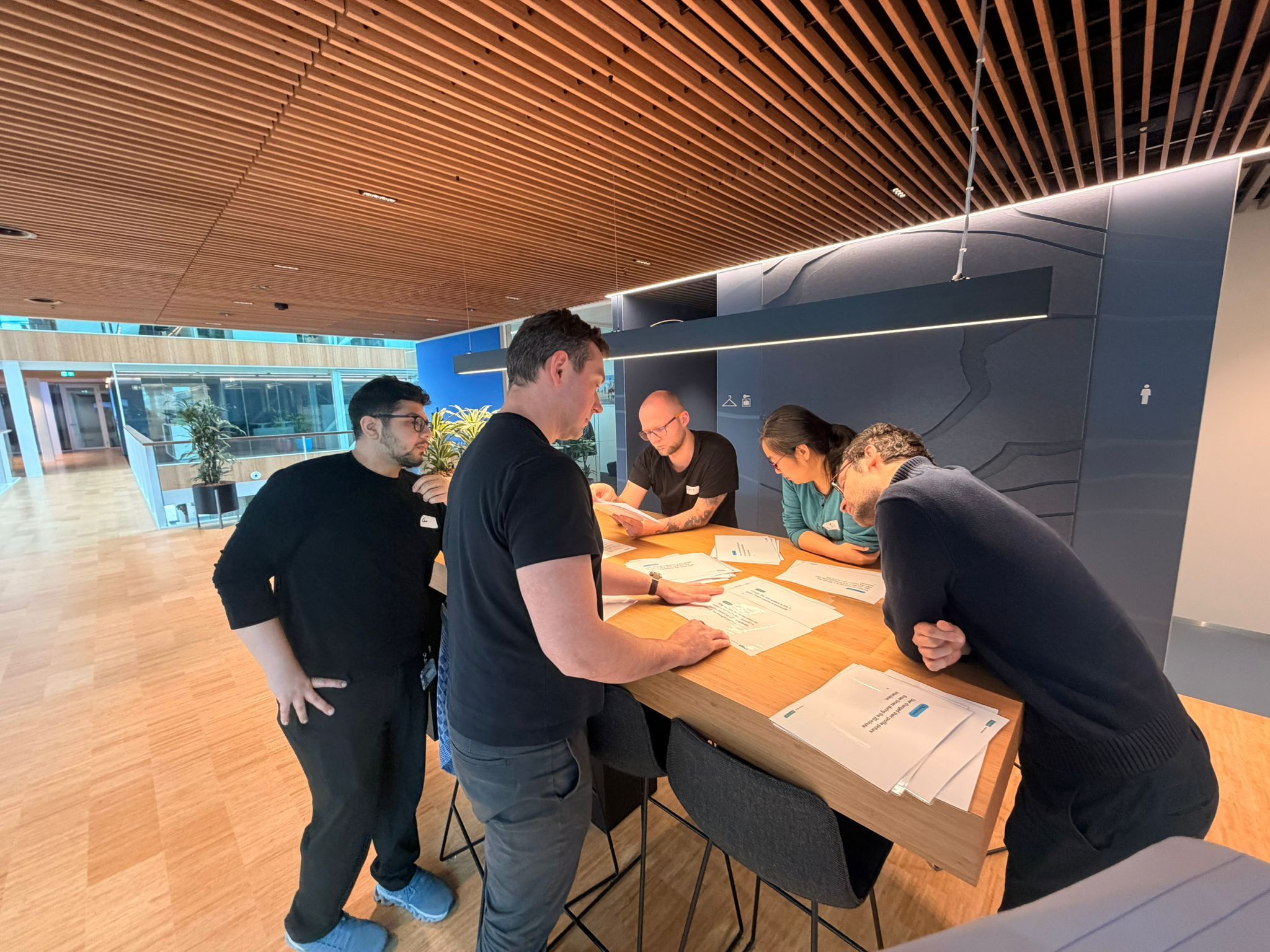 Five people around a high table in an open office, printed research sheets spread across it. Several are leaning in over the same sheets, one is holding a page up to read from it, and one stands back with hands on hips watching the group work.